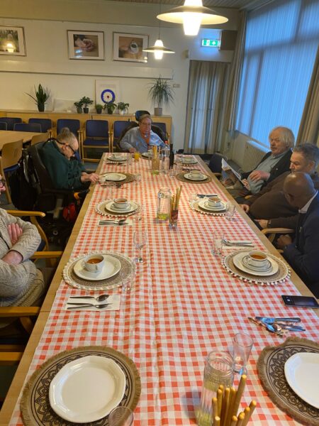 Een groep van zeven personen zit rond een rechthoekige tafel met een rood-wit geblokt tafelkleed, gedekt met borden en kopjes, in een kamer met stoelen en planten.